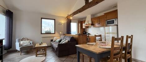 A cozy living room with a sofa, a small table, and a dining table with chairs. There is a kitchen area with wooden cabinets and appliances.
