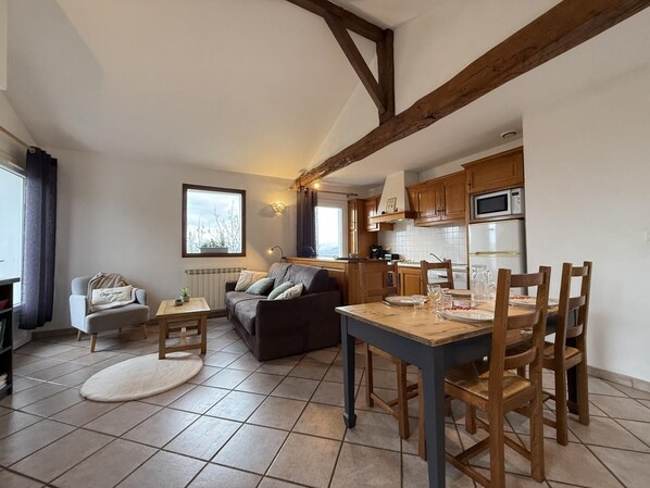 A cozy living room with a sofa, a small table, and a dining table with chairs. There is a kitchen area with wooden cabinets and appliances.