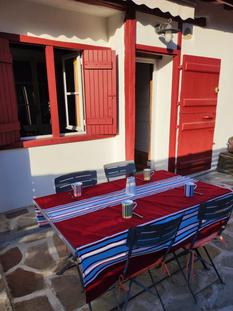 Un'area pranzo all'aperto con un tavolo imbandito per quattro persone, circondata da pavimento in pietra e finestre con infissi rossi.