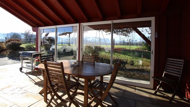 An outdoor patio with a round wooden table and chairs, a grill, and a view of a mountainous landscape through large sliding glass doors. - Haizalde - Saint-jean-le-vieux (SAINT-JEAN-LE-VIEUX)