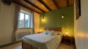 A bedroom with a wooden bed, a nightstand with a lamp, a window with curtains, and a green wall. - Haizalde - Saint-jean-le-vieux (SAINT-JEAN-LE-VIEUX)