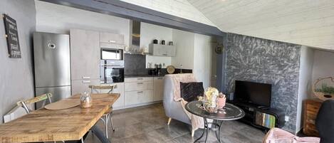 Una cocina moderna con electrodomésticos integrados, una zona de comedor con mesa y sillas de madera, y un rincón de estar con sofá y televisor.