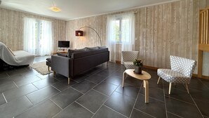 Un salon moderne comprenant un canapé, deux fauteuils à motifs, une petite table avec une plante, un téléviseur et un panneau mural en bois.