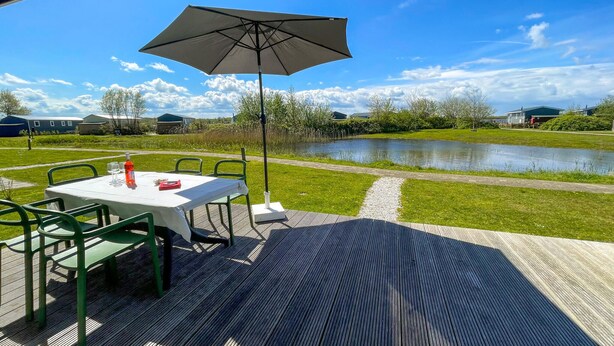 Un patio al aire libre con mesa y sillas, una sombrilla grande, y vista a un estanque y follaje circundante.