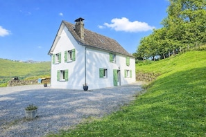 Una casa blanca con contraventanas verdes, un camino de grava y un muro de piedra al fondo.