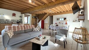 Een ruime woonkamer met een bank, salontafel en een eettafel voor vier personen. Een trap leidt naar een hoger gelegen verdieping, en een televisie hangt aan de muur.