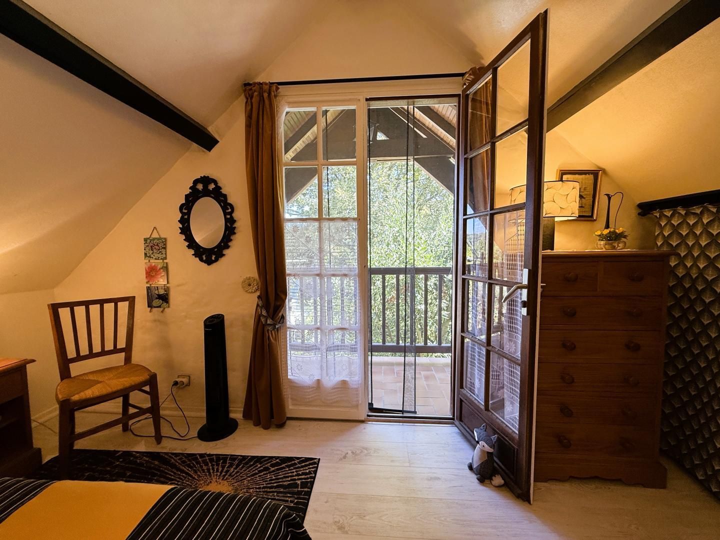 A room with a large sliding glass door leading to a balcony, a wooden chair, a small table with a lamp, and a dresser with a patterned cover.