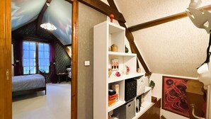 A room with a sloped ceiling, a bed, a white shelving unit with various items, and a painting on the wall.