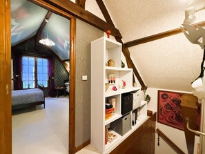 A room with a sloped ceiling, a bed, a white shelving unit with various items, and a painting on the wall. - Cauhape - Auga - Gîte (AUGA)