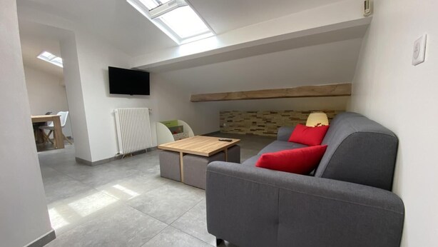 A modern living room with a grey sofa, a wooden coffee table, and a TV mounted on the wall. There is a window with a view of the outside. - Mendi Bichta - Halsou (HALSOU)