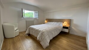 A bedroom with a bed, bedside lamps, a chair, a radiator, and a window with a view of greenery. - Mendi Bichta - Halsou (HALSOU)