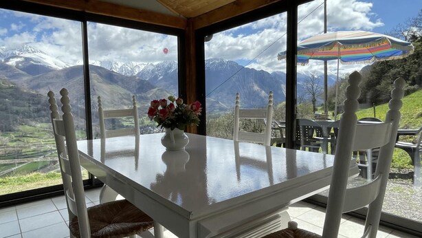 A dining area with a table and chairs, a vase with flowers, and a view of mountains through large windows.