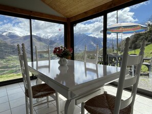 A dining area with a table and chairs, a vase with flowers, and a view of mountains through large windows.
