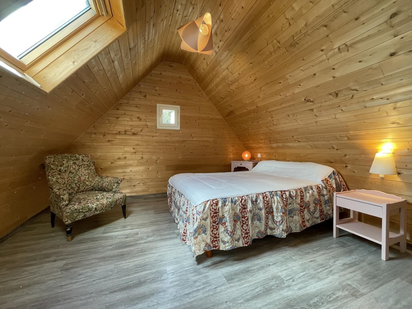 A cozy attic bedroom with a sloped ceiling, a bed with a floral bedspread, a chair, a bedside table, and a lamp.