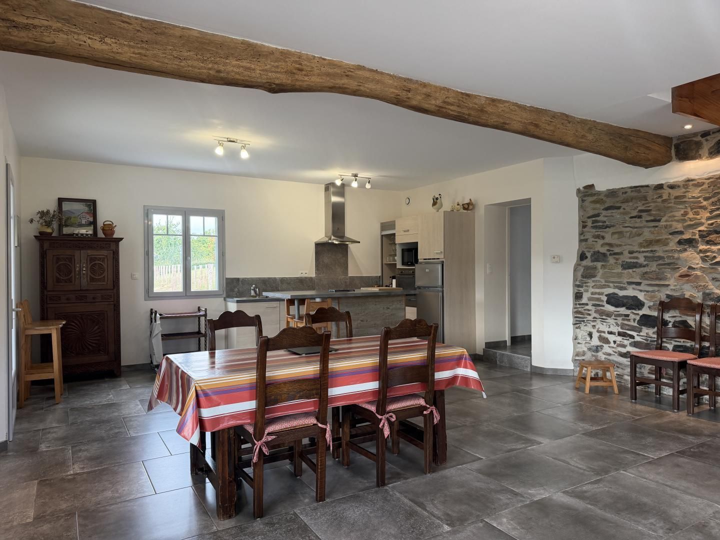 Un comedor espacioso con una larga mesa de madera y sillas, una cocina al fondo y una pared de piedra.
