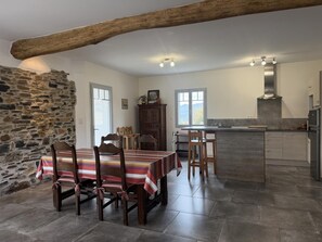 Un coin repas avec une table dressée pour quatre personnes, une cuisine avec un mur en pierre et un grand plafond avec une poutre en bois.