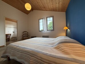Une chambre avec un grand lit, un plafond en bois, deux fenêtres, une lampe de chevet et une vue sur l’extérieur.