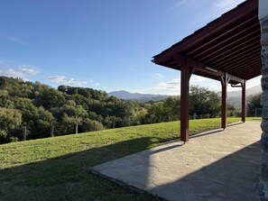 A stone patio with a pergola overlooking a grassy field and forested hills.