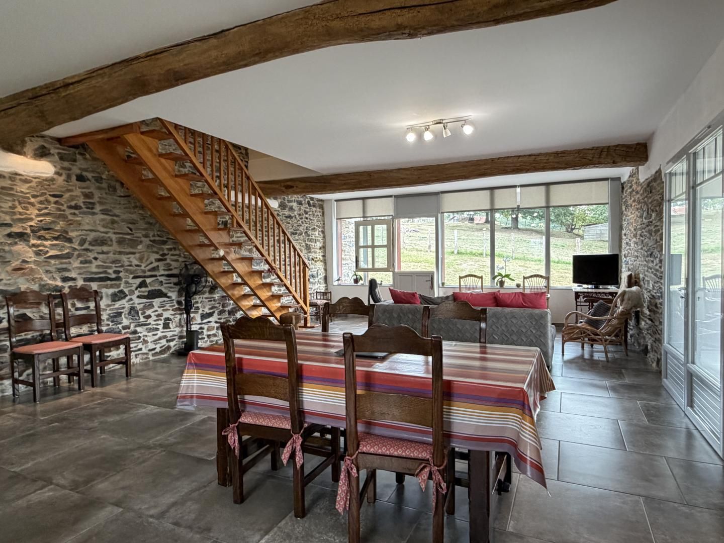 A spacious dining area with a wooden staircase, stone walls, and large windows offering a view of the outdoors.