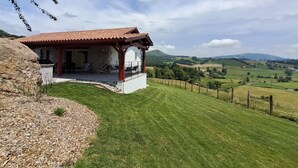 Ein Steinhäuschen mit rotem Ziegeldach und einer überdachten Terrasse. Es liegt auf einem grasbewachsen Hügel mit Blick auf sanfte Hügellandschaft und einen klaren Himmel.