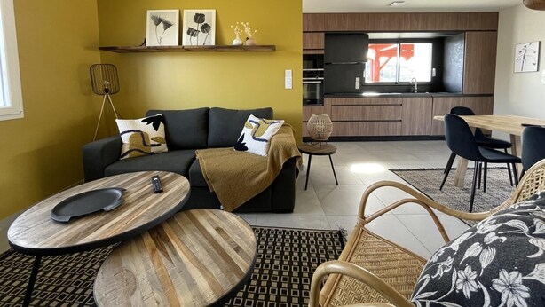Ein modernes Wohnzimmer mit Sofa, Couchtisch und Essbereich. Im Hintergrund ist eine Küche mit einem großen Fenster zu sehen.