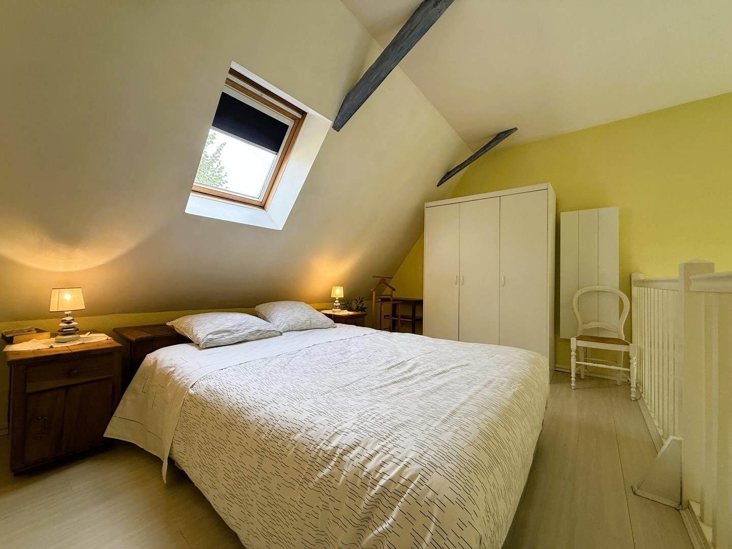 Un dormitorio con techo inclinado, un tragaluz, una cama con colcha estampada, una mesita de noche con lámpara, un armario y una silla.