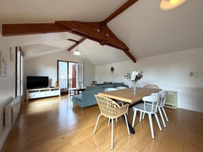A spacious living area with a wooden ceiling, a dining table with chairs, a sofa, and a television.