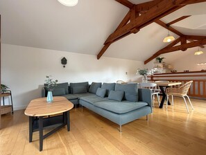 A spacious loft with a high ceiling, exposed wooden beams, a sectional sofa, a round wooden table, and a dining area with wicker chairs.