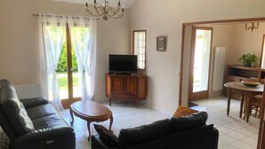 En stue med sofa, et lille bord, et fjernsyn og et skab. Der er en dør til en spiseplads med et bord og stole.