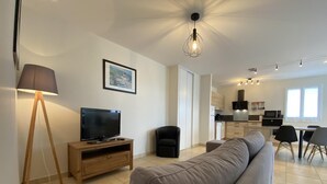 A modern living room with a flat-screen TV, a wooden TV stand, a grey sofa, a floor lamp, and a dining area with a table and chairs. - Maliania - Arbonne - Gîte (ARBONNE)