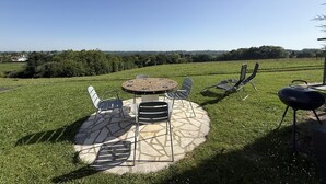 An outdoor patio with a round table and chairs, a barbecue grill, and a view of a grassy field and distant trees. - Maliania - Arbonne - Gîte (ARBONNE)
