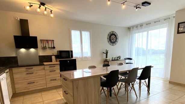 A modern kitchen with a central island, dining table, and chairs. There are windows with curtains, a clock on the wall, and a flat-screen TV mounted above the counter. - Maliania - Arbonne - Gîte (ARBONNE)