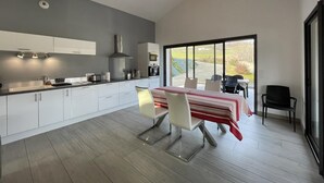 A modern kitchen with white cabinets, a dining area with a red-striped tablecloth, and a large window offering a view of the outdoors. - Buztingorria - Macaye (MACAYE)