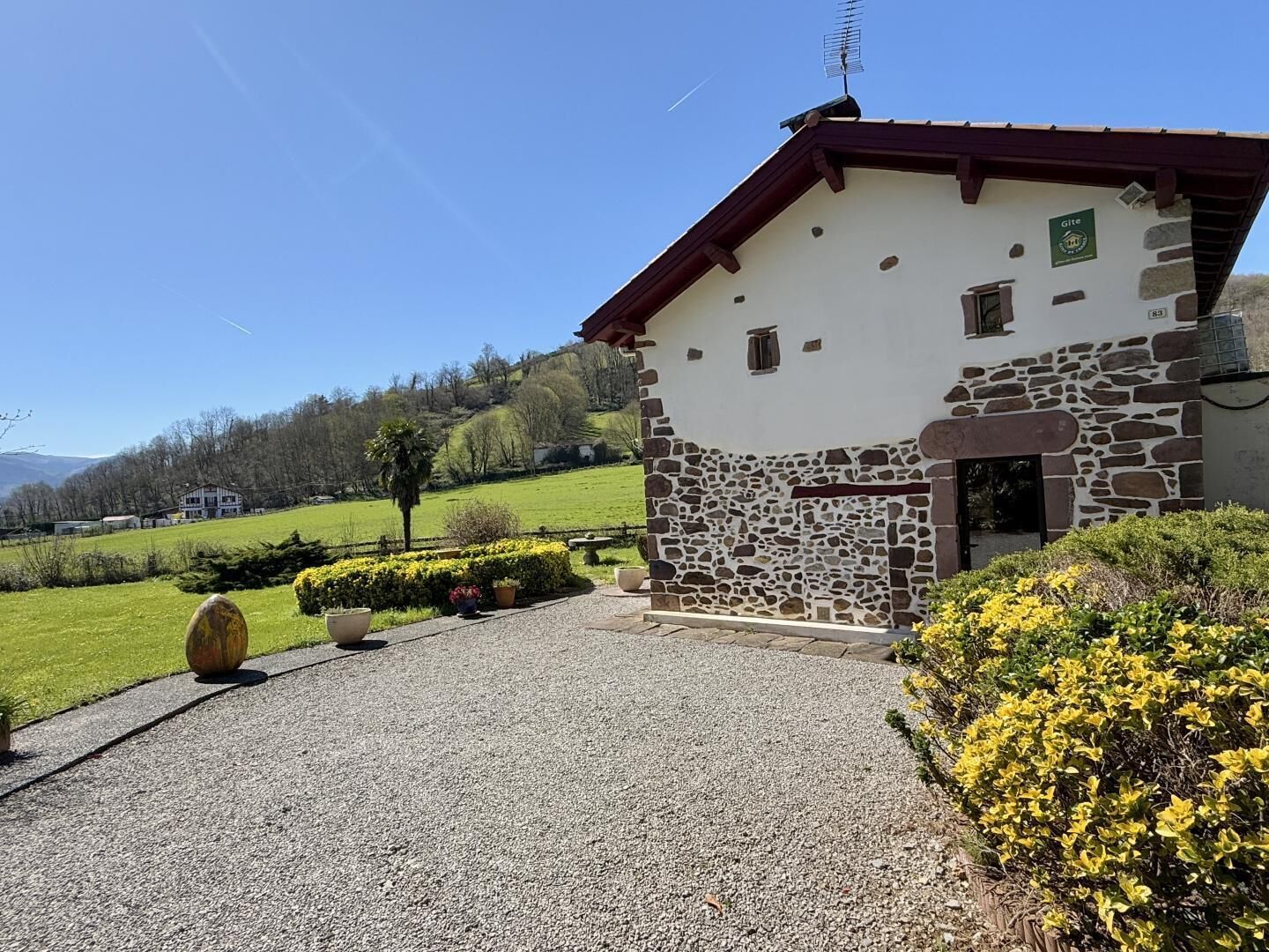 A stone building with a sloped roof, a gravel driveway, and a well-maintained garden with yellow flowers.