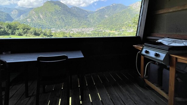 Une cabane rustique avec un intérieur en bois, une table à manger, des chaises et un réchaud à gaz. Une grande fenêtre offre une vue sur des montagnes verdoyantes et une vallée.