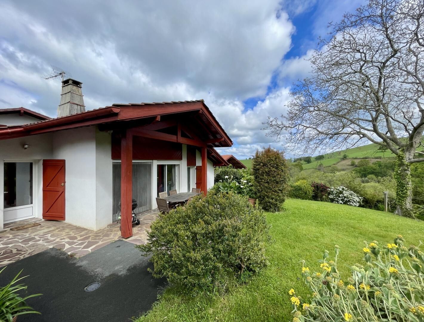 A house with a red roof, a stone patio, and a garden with flowers and a tree.