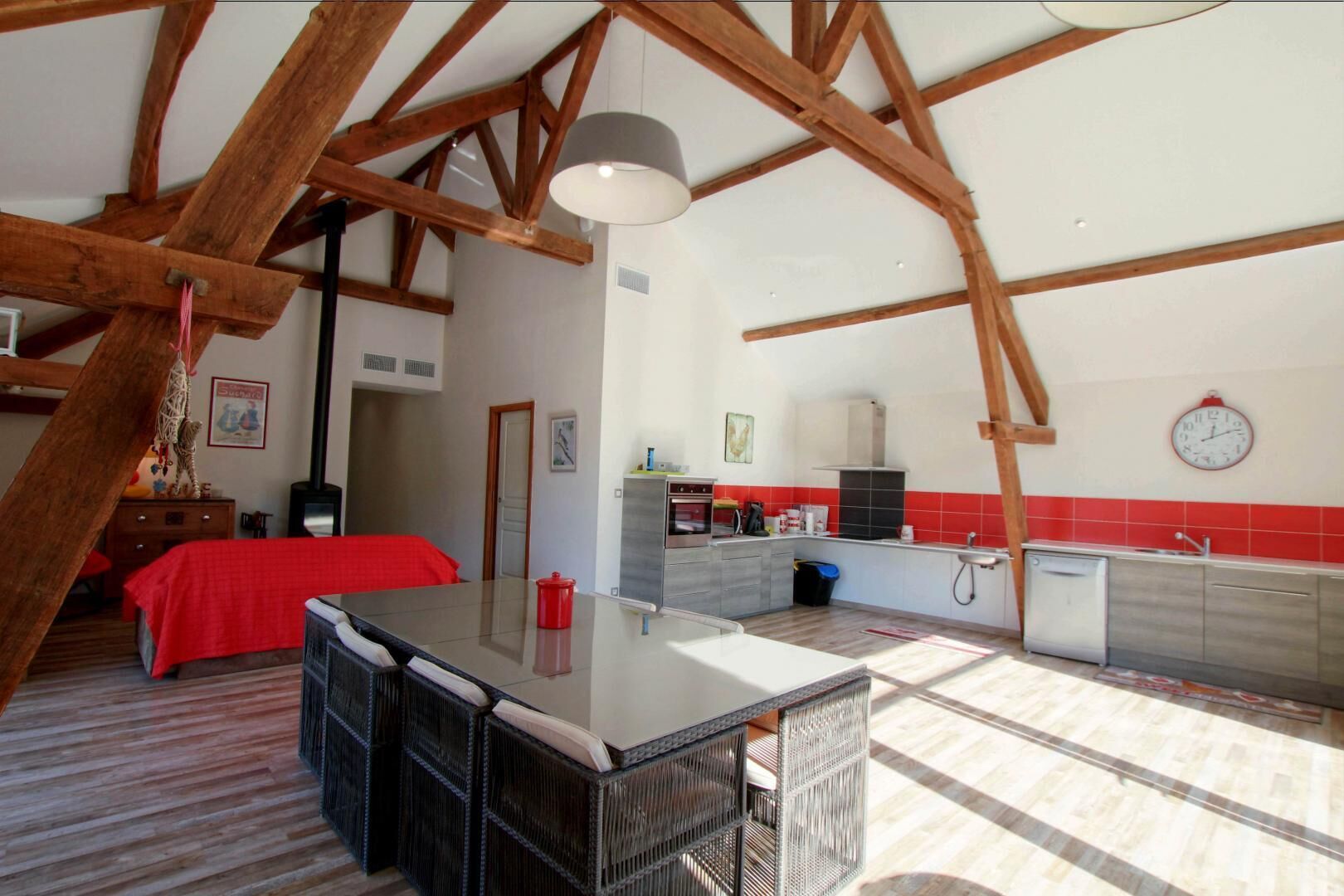 A spacious loft with a high ceiling, exposed wooden beams, a kitchenette, a dining area, and a bedroom with a red bedspread.