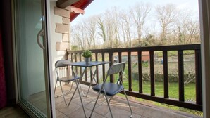 A balcony with a table and two chairs, a potted plant, and a view of a yard with trees and a red-roofed building. - Xearbaita - Urrugne - Gîte (URRUGNE)