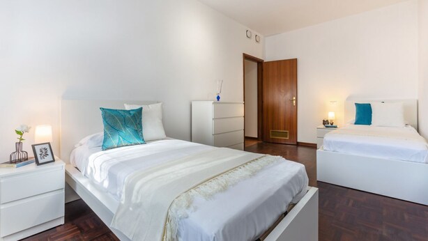 A bedroom with a large bed, a smaller bed, a nightstand with a lamp, a dresser, and a wooden door. - GuestHost - Pontelagoscuro Huge & Residential Apartment (Ferrara)