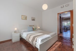 A bedroom with a wooden floor, a bed with a white headboard, two bedside tables with lamps, and a door leading to a dining area.