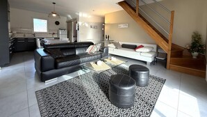 Un salon moderne avec un canapé en cuir noir, un canapé blanc et une table basse en verre. Un escalier avec des rampes en bois mène à un niveau supérieur.