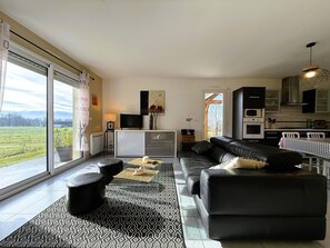 A modern living room with a black leather sofa, a dining table with a checkered tablecloth, a kitchen area with a microwave and oven, and a large window with a view of a green field.