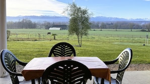 Une terrasse couverte, avec une table et des chaises, donnant sur un champ verdoyant et offrant une vue sur les montagnes en arrière-plan.