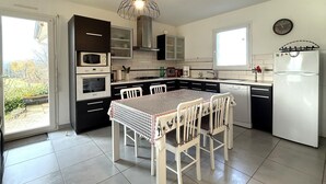 Une cuisine moderne avec un coin repas, comprenant une nappe à carreaux, des chaises blanches, un luminaire suspendu et un réfrigérateur.