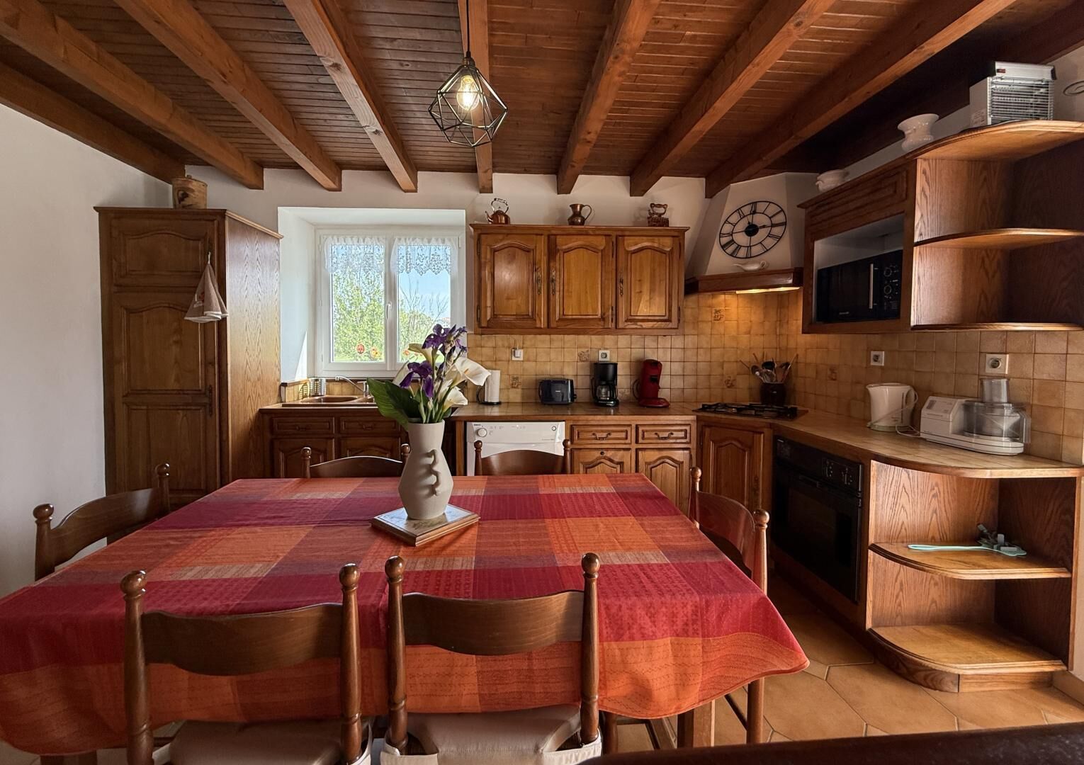A kitchen with wooden cabinets, a dining table with chairs, a vase with flowers, and a clock on the wall.