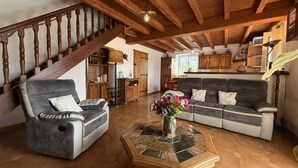 Een ruime woonkamer met een bank, fauteuil en een salontafel met een bloevas. Een houten trap en een keuken zijn zichtbaar op de achtergrond.