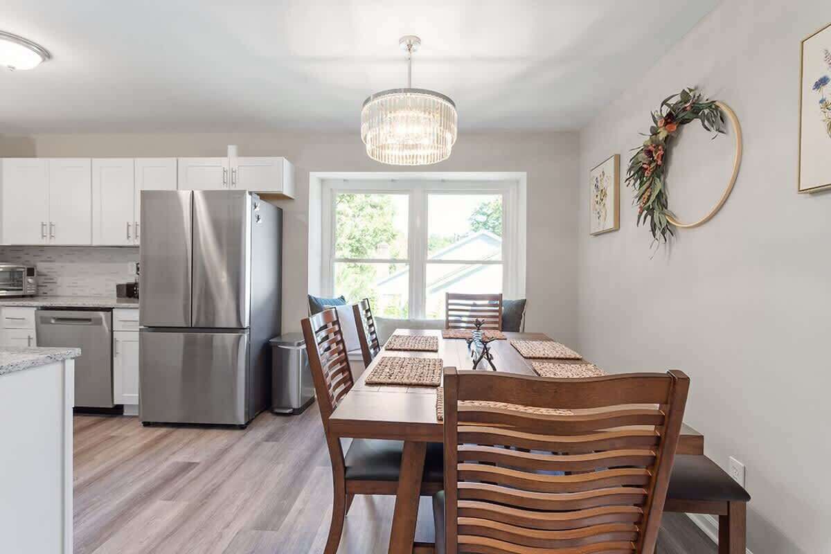 A modern kitchen with white cabinets, a stainless steel refrigerator, wooden chairs, and a chandelier.