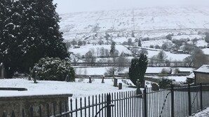 A snow-covered cemetery with a black metal fence, a large evergreen tree, and a distant view of a hilly landscape. - Holiday Cottage In The Village Of Dent In The Yorkshire Dales - Dog friendly (Dent)