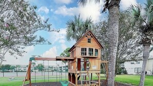 A wooden playhouse with a swing set and climbing area, surrounded by trees and a fence. - Prime Gulf Highlands Property: 0.4 Mile to Private Beach & 11 Pools! (Panama City Beach)