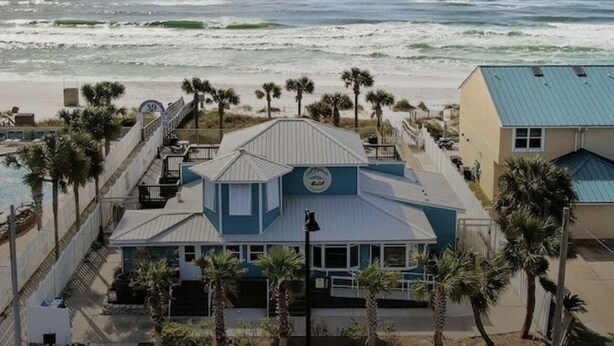 A beachfront property with a prominent building featuring a blue roof and a balcony overlooking the ocean. Palm trees line the property, and the beach is visible in the background. - Prime Gulf Highlands Property: 0.4 Mile to Private Beach & 11 Pools! (Panama City Beach)
